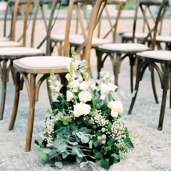 Stolar till bröllopsgäster står uppradade med ett vitt blomsterarrangemang framför.